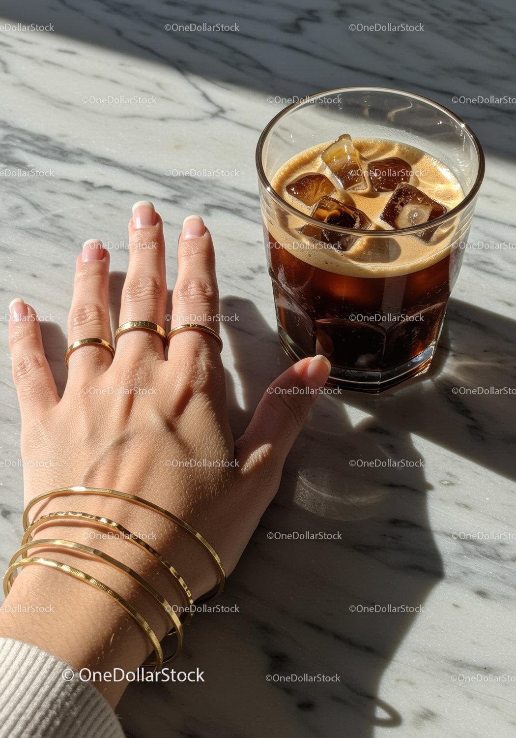 Hand with Gold Jewelry and Glass of Iced Coffee on Marble