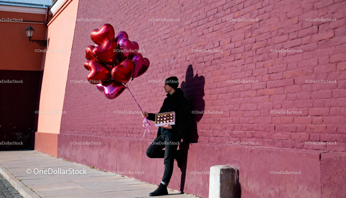 Man Holding Heart Balloons and Chocolates by Brick Wall