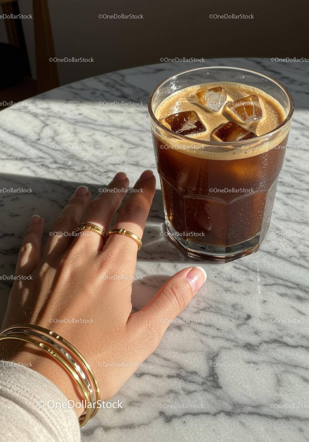 Hand with gold jewelry next to iced coffee on marble table