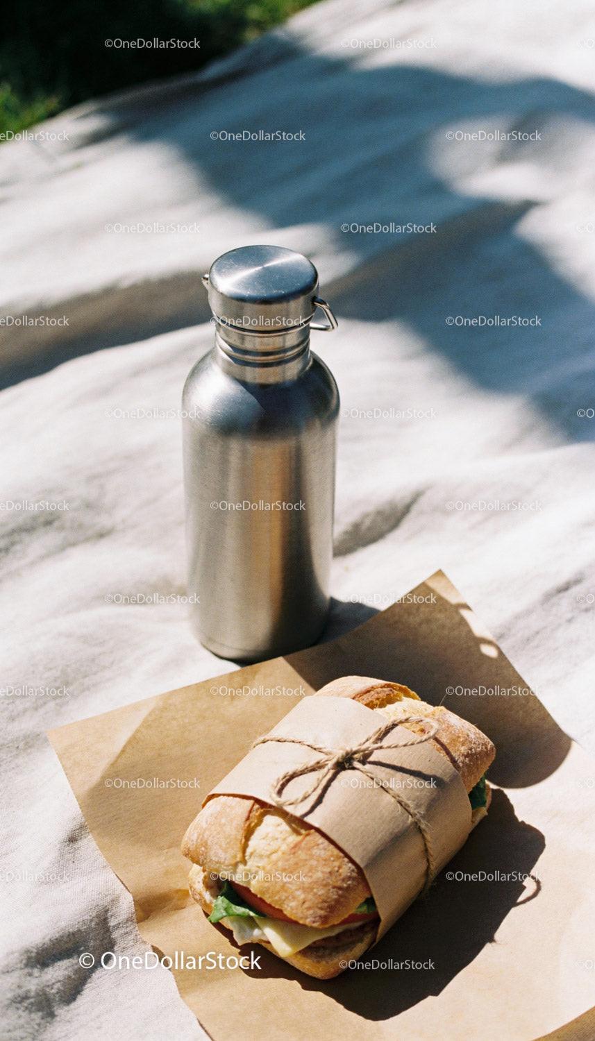 Rustic Sandwich Wrapped with Paper Next to Metal Bottle