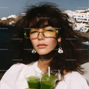 Stylish Woman with Yellow Sunglasses Holding Green Juice by the Sea
