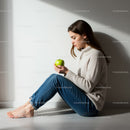 Young woman sitting by wall holding green apple