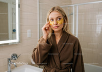 Woman in Brown Bathrobe Applying Eye Mask for Skincare Treatment