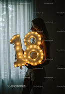 Young woman holding illuminated 18 birthday balloon indoors