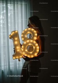 Young woman holding illuminated 18 birthday balloon indoors