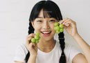 Smiling Asian Woman Holding Green Grapes in Bright Indoor Setting