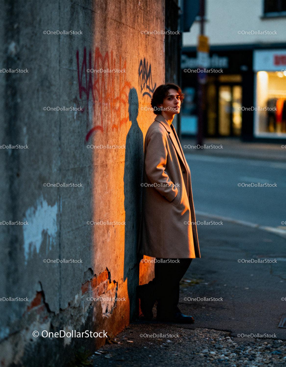 Young man in beige coat leaning on urban wall at sunset