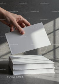 Hand Holding a Blank White Book Above Stacked Books