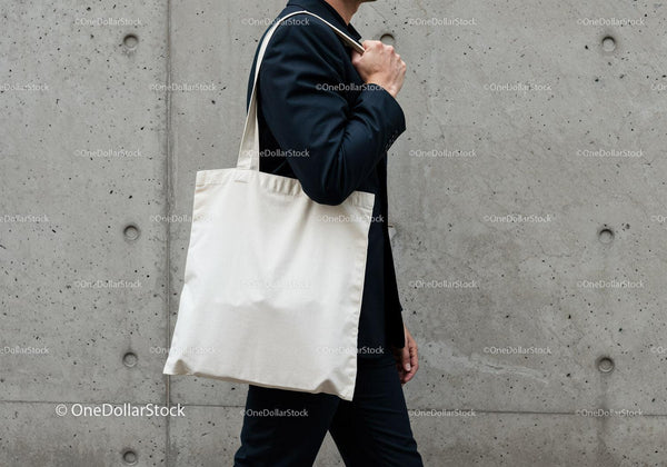 Person carrying blank canvas tote bag walking by concrete wall JPG Mockup Image