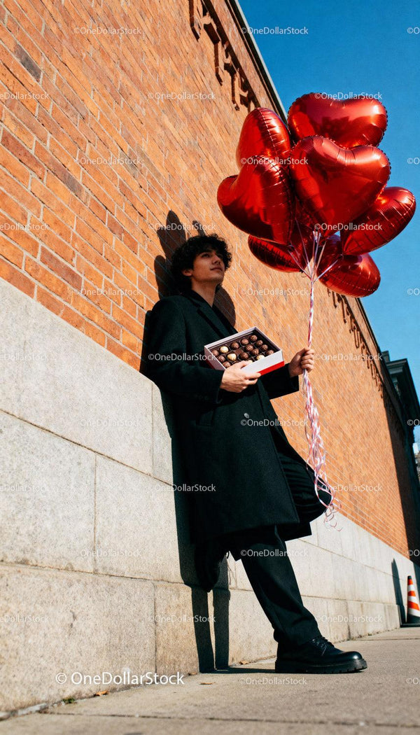 Young man holding heart balloons and chocolates outdoors