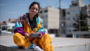 Young Woman Sitting on Urban Street in Colorful Fashion Smiling at Tablet