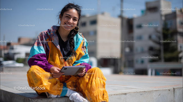 Young Woman Sitting on Urban Street in Colorful Fashion Smiling at Tablet