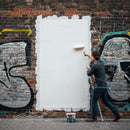 Man painting over graffiti with white roller on brick wall
