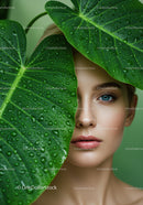 Close-up of Woman with Blue Eyes Behind Large Tropical Green Leaves with Water Droplets