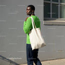Young Man in Green Sweater with Tote Bag Standing against Gray Wall