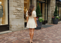 Fashionable Woman in Beige Dress Walking Past Storefront with Flower Pots
