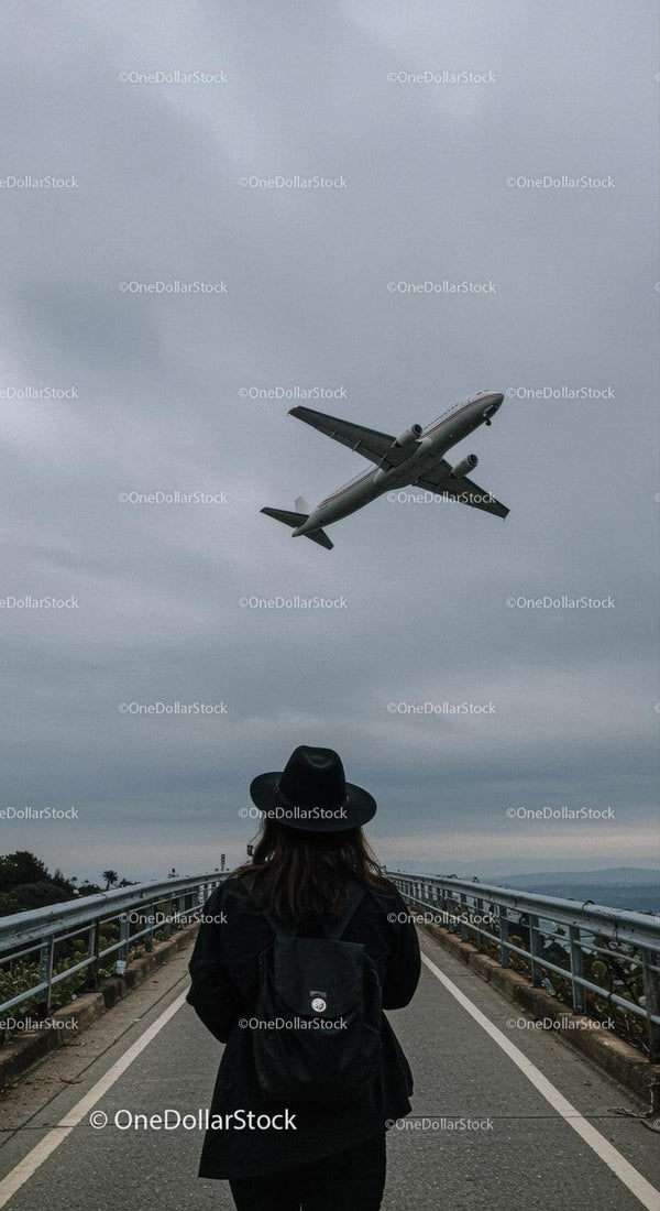 Woman in Black Hat Watching Airplane Flying Over Bridge in Urban Environment