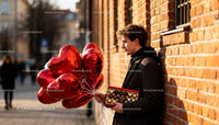 Young man holding chocolates and heart balloons outdoors