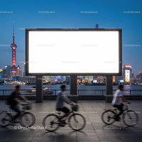 Evening cyclists passing large empty billboard by city river