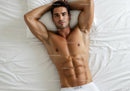 Relaxed muscular man lying on white bed with arms behind head