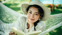 Smiling Young Woman Relaxing in Hammock Reading Book in Garden