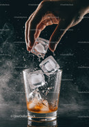 Hand adding ice cubes to amber drink in glass tumbler