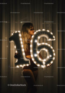 Young woman holding illuminated 16-number balloons indoors