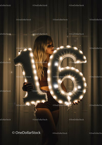 Young woman holding illuminated 16-number balloons indoors