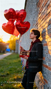 Young man holding chocolates and heart balloons outdoors
