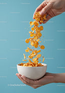 Hand pouring corn flakes into white bowl on blue