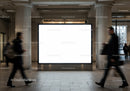 Busy commuters walking past large blank billboard indoors