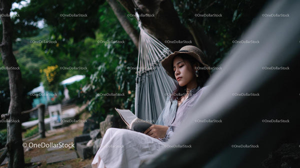 Young Woman Relaxing in Hammock Reading Book in Lush Green Park