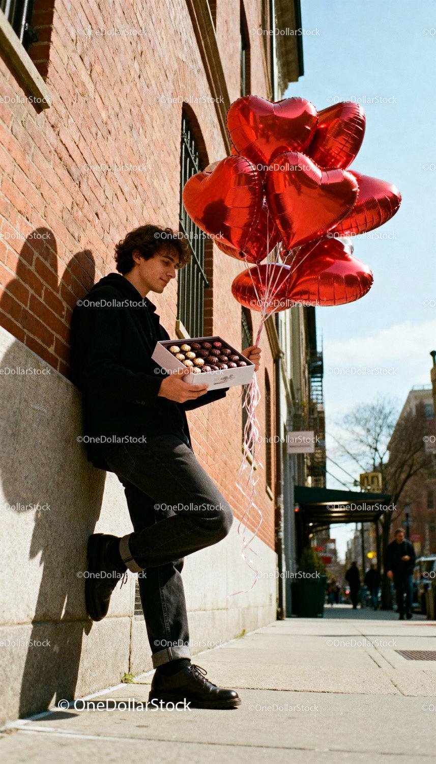 Young man holding heart balloons and chocolates outdoors