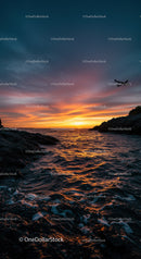 Serene Sunset Over Ocean with Rocky Shore and Airplane in Sky