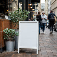Blank white sandwich board on urban sidewalk with greenery