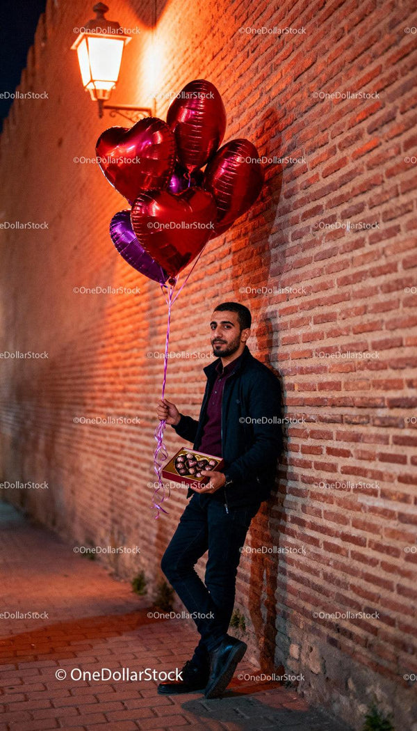 Man with heart balloons and chocolates near brick wall