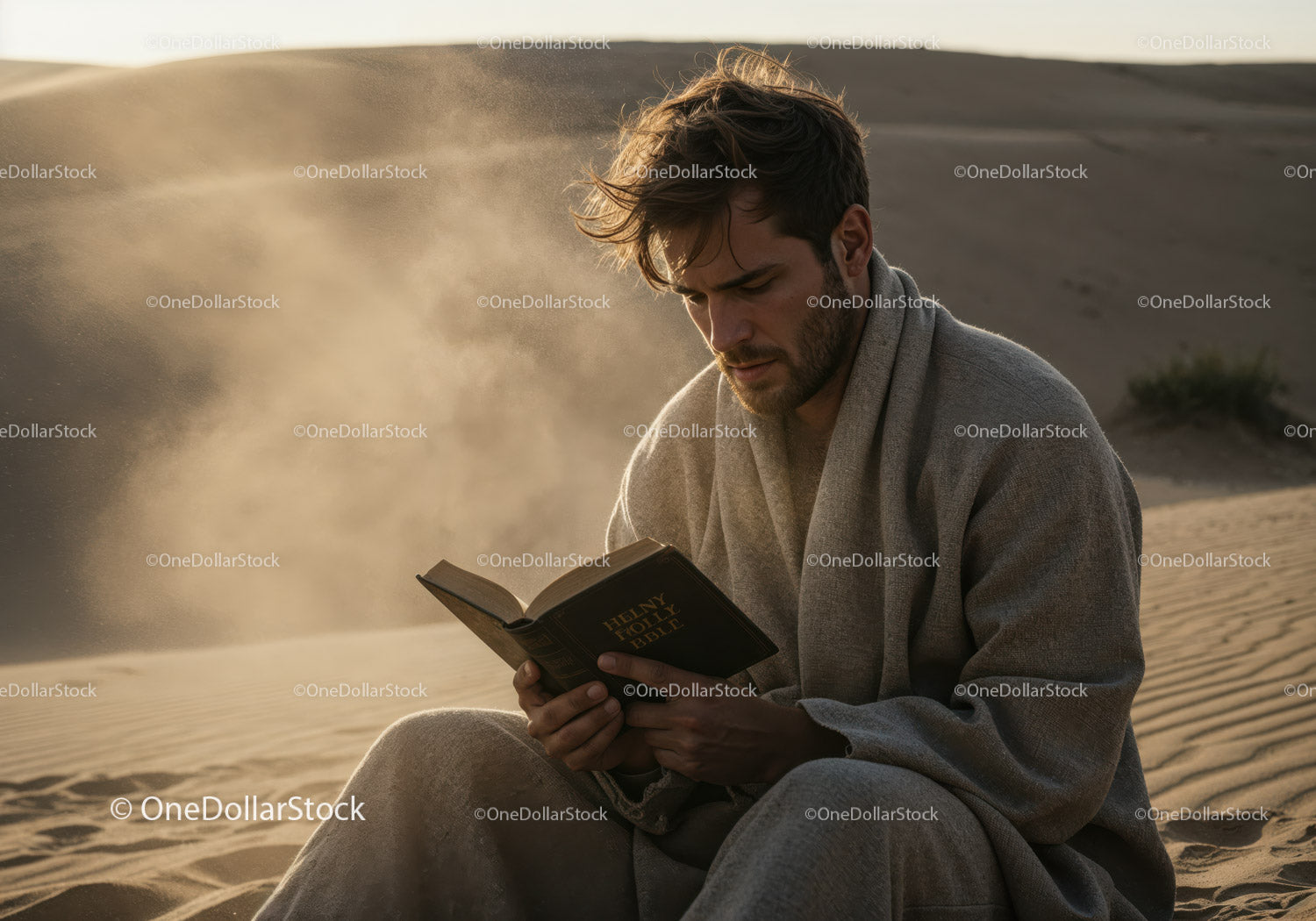 Young Man Reading Bible in Desert Surrounded by Sand Dunes at Sunset