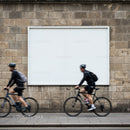 Two cyclists riding past blank urban billboard