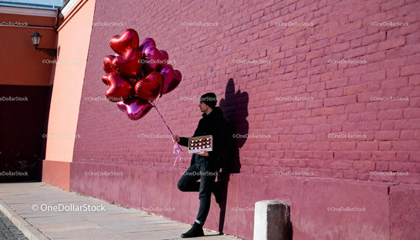 Man Holding Heart Balloons and Chocolates by Brick Wall