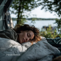 Peaceful outdoor scene of a woman sleeping by a lakeside beneath lush green trees