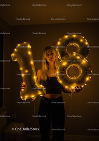 Young woman holding illuminated number 18 balloons indoors