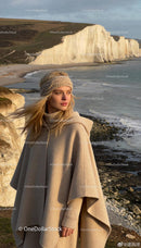 Woman in warm beige cape and headband by coastline cliffs