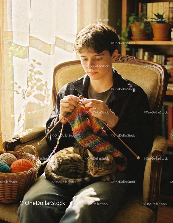 Young man knitting colorful scarf with cat on lap