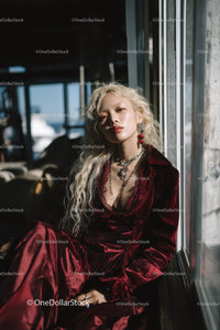 Elegant blonde woman in red velvet suit sitting by window with sunlight
