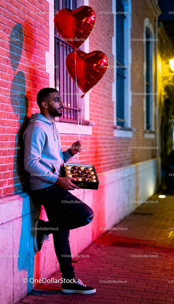 Young man holding chocolates and heart-shaped balloons outdoors
