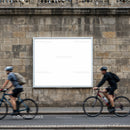 Two cyclists riding past blank urban billboard on stone wall