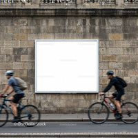 Two cyclists riding past blank urban billboard on stone wall